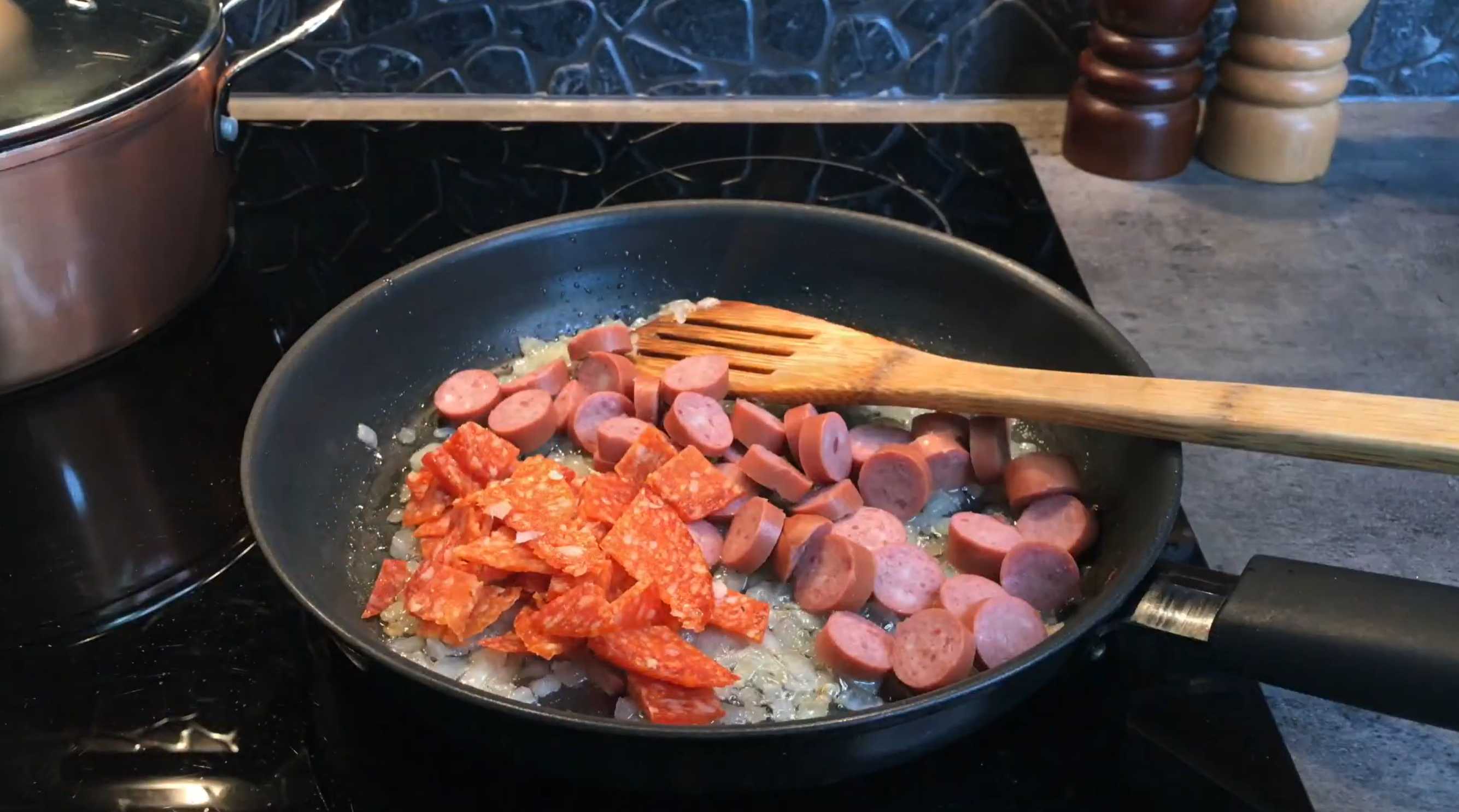 Далее нарезаем сосиски и охотничьи колбаски. Добавляем нарезанные колбасные изделия к луку, обжариваем, постоянно помешивая. Солянка на сковороде-Шаг 3